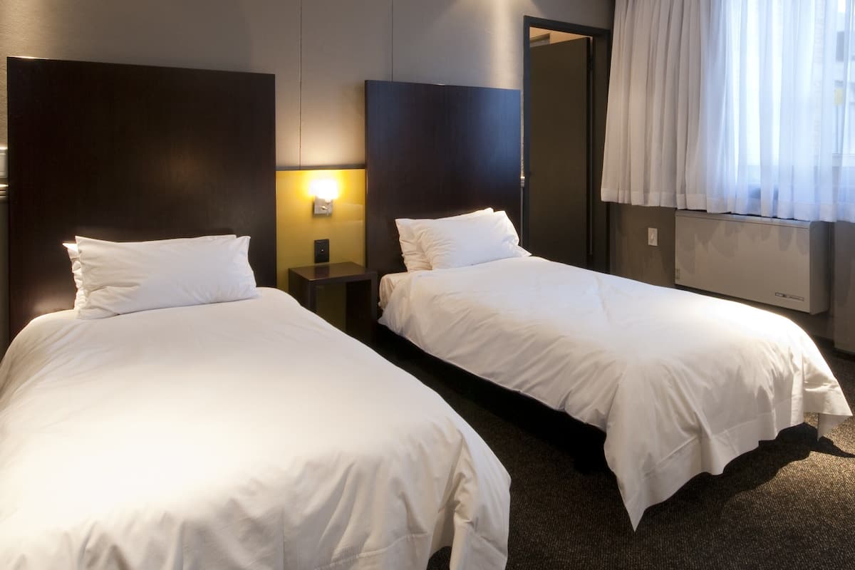 Une chambre d’hôtel avec deux lits simples, une table de chevet avec une lampe, un grand miroir et une fenêtre avec des rideaux.