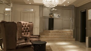 Un lobby lujoso con un sillón de cuero capitonado, un otoman redondo, una araña y una escalera que conduce al centro de negocios.
