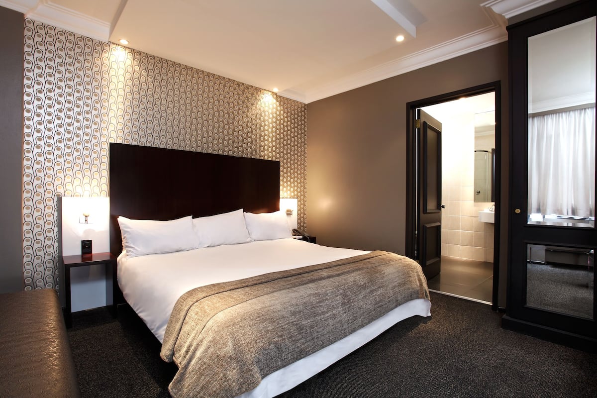 Une chambre d’hôtel avec un grand lit, une table de chevet avec une lampe, un papier peint à motifs et une porte donnant sur une salle de bain.