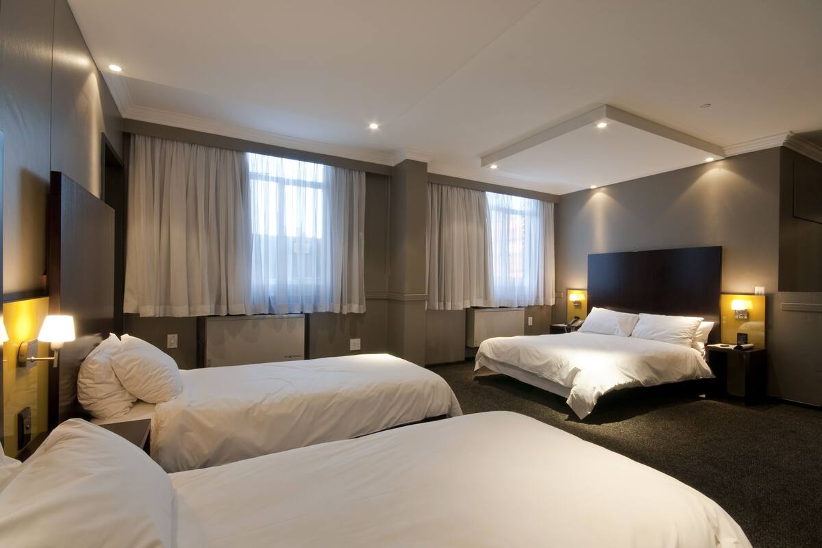 Une chambre d’hôtel avec deux lits simples, une grande fenêtre avec des rideaux, un téléviseur à écran plat et une table de chevet avec une lampe.