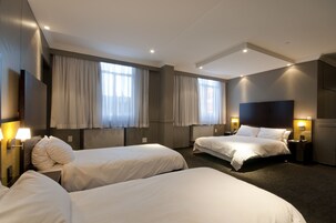 Habitación de hotel con dos camas individuales, un ventanal con cortinas, un televisor de pantalla plana y una mesita de noche con lámpara.