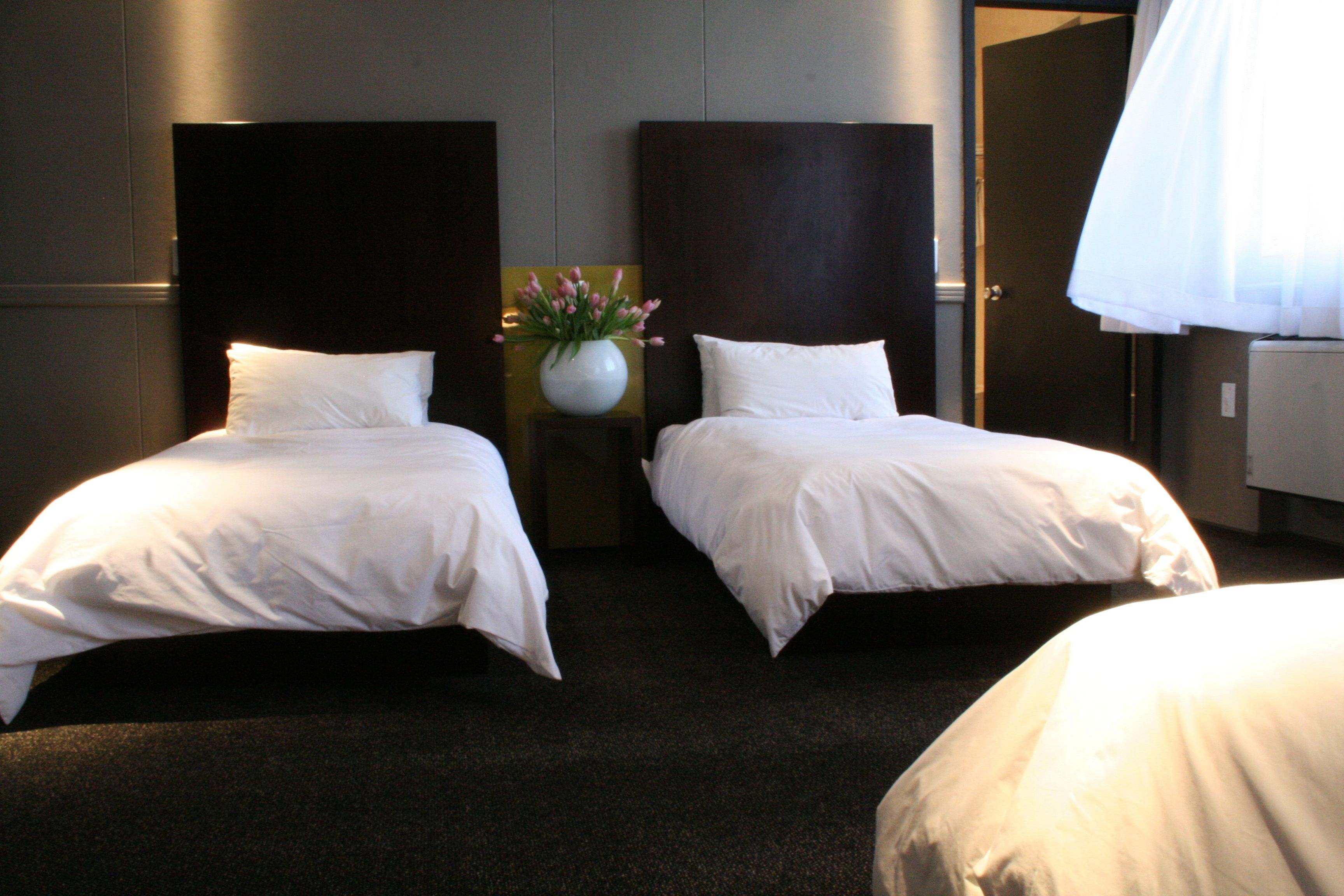 A hotel room with two single beds, a vase with flowers on a nightstand, and a large window with curtains.
