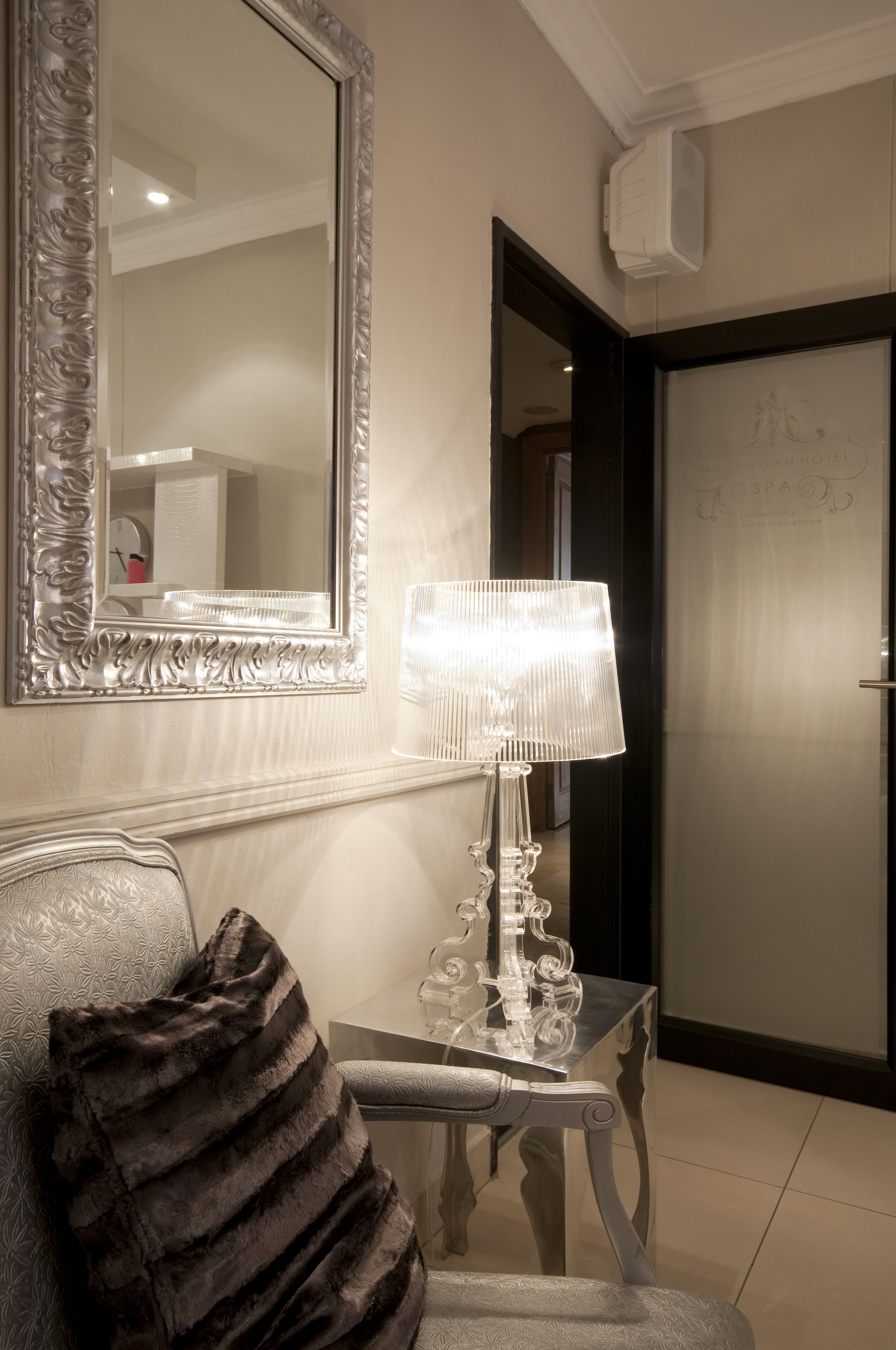A hotel room with a large ornate mirror, a table with a lamp, and a plush chair with a dark throw pillow.