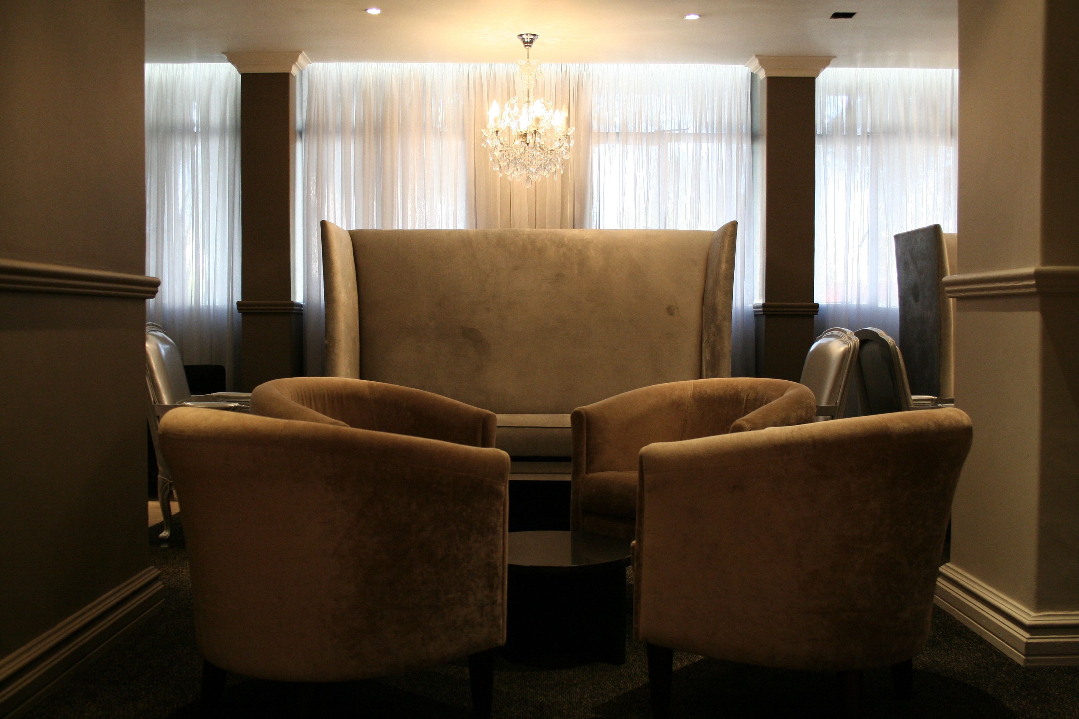 A room with a large armchair and two smaller armchairs facing a large window with sheer curtains. A chandelier hangs from the ceiling.