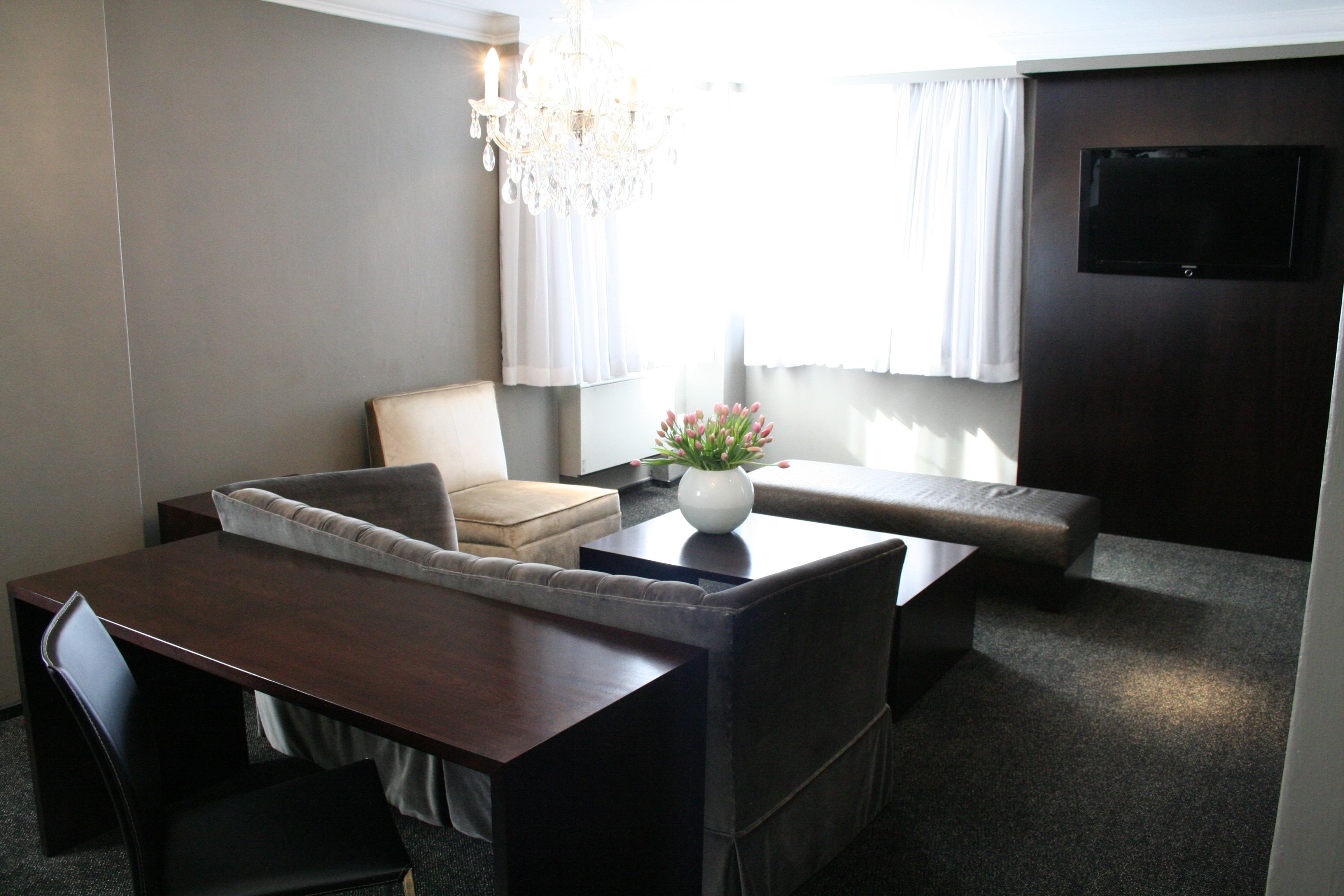 A modern hotel room with a dining area, a sofa, a loveseat, a small table with a vase of flowers, and a TV mounted on the wall.