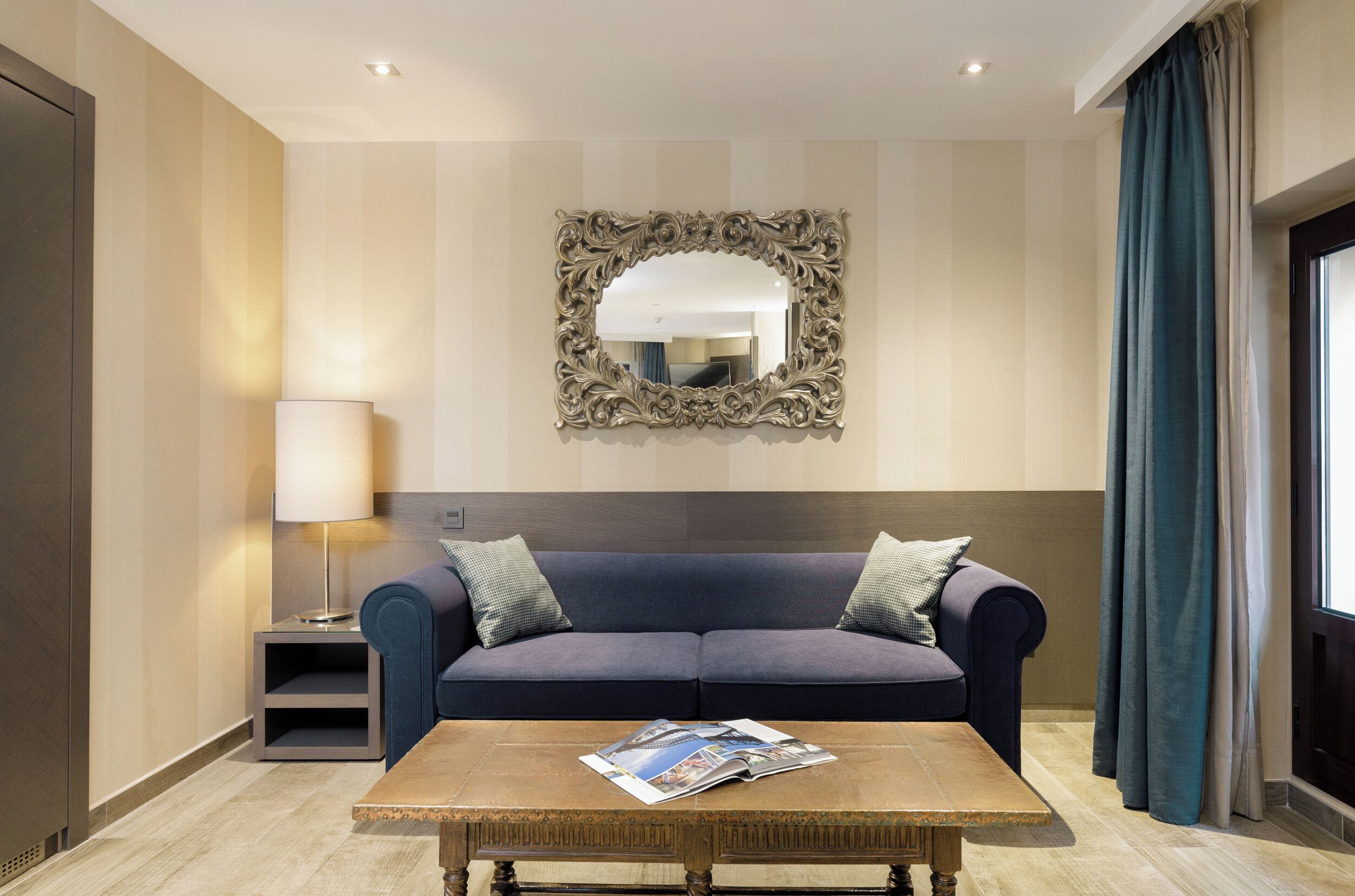Una habitación de hotel moderna con un sofá azul oscuro, una mesa de centro de madera con una revista, un espejo decorativo y una lámpara.
