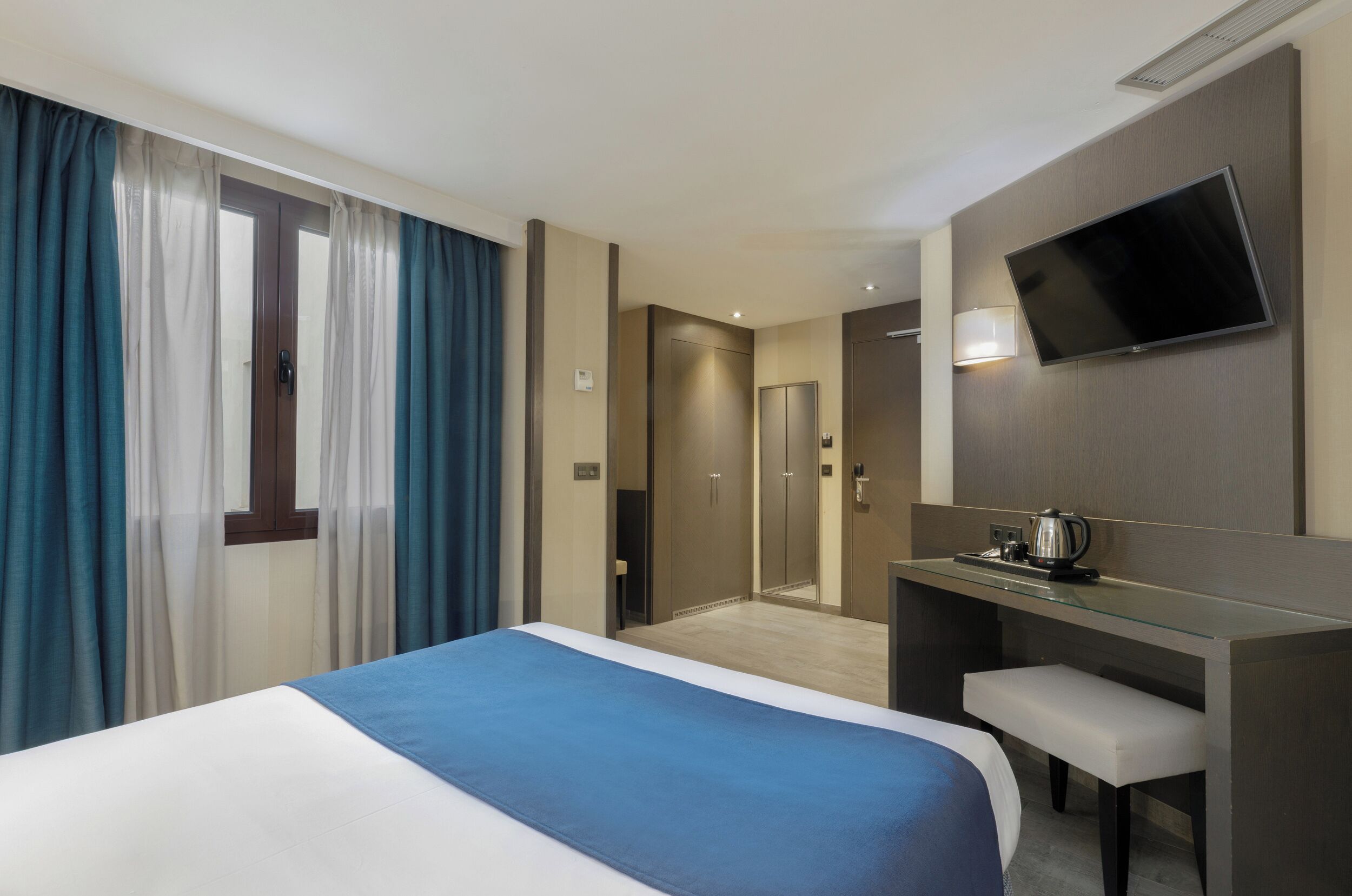 A hotel room with a bed, a desk with a chair, a television mounted on the wall, and a bathroom visible through a door.