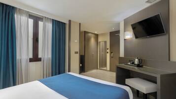 A hotel room with a bed, a desk with a chair, a television mounted on the wall, and a bathroom visible through a door.