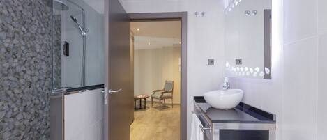 A modern bathroom with a glass shower enclosure, a white sink, and a chair in the adjacent room.