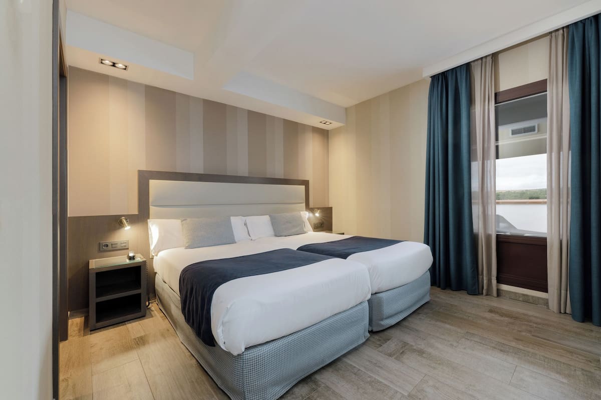 Cama doble con cabecera, mesitas de noche y vistas al exterior a través de una ventana con cortinas.