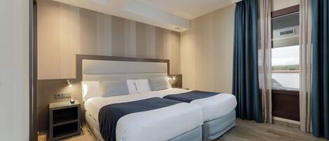 Cama doble con cabecera, mesitas de noche y vistas al exterior a través de una ventana con cortinas.