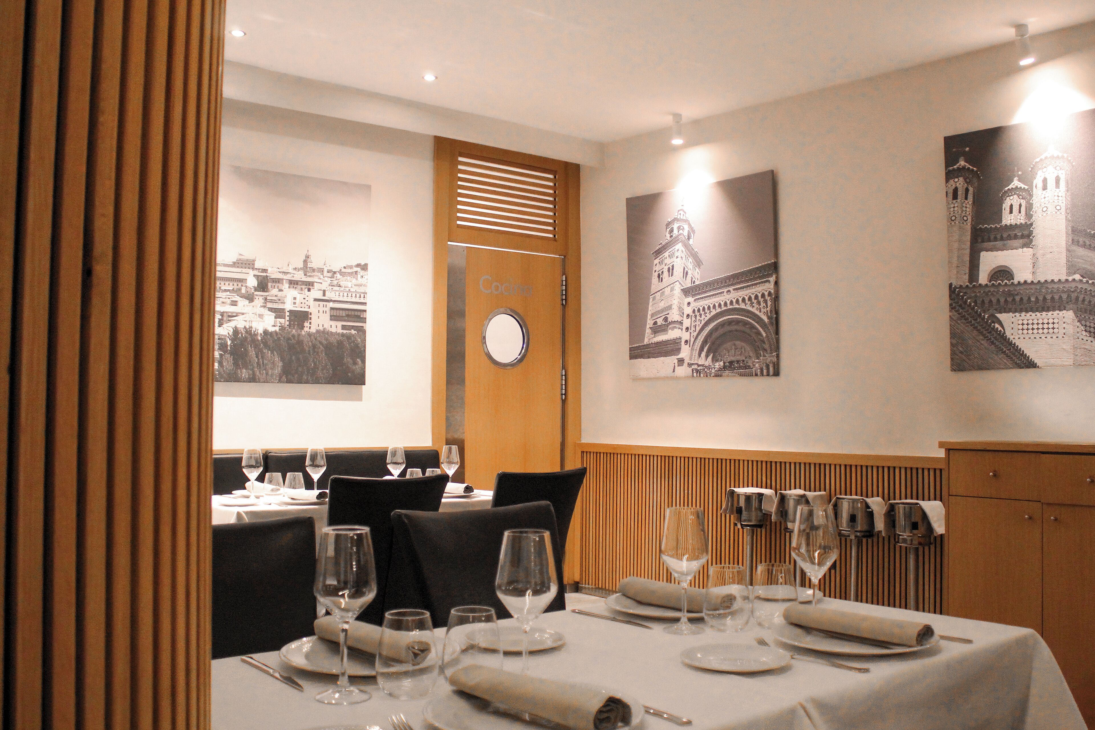 Un comedor con mesas dispuestas para una comida, una puerta de madera con un ventanal circular y cuadros en la pared.
