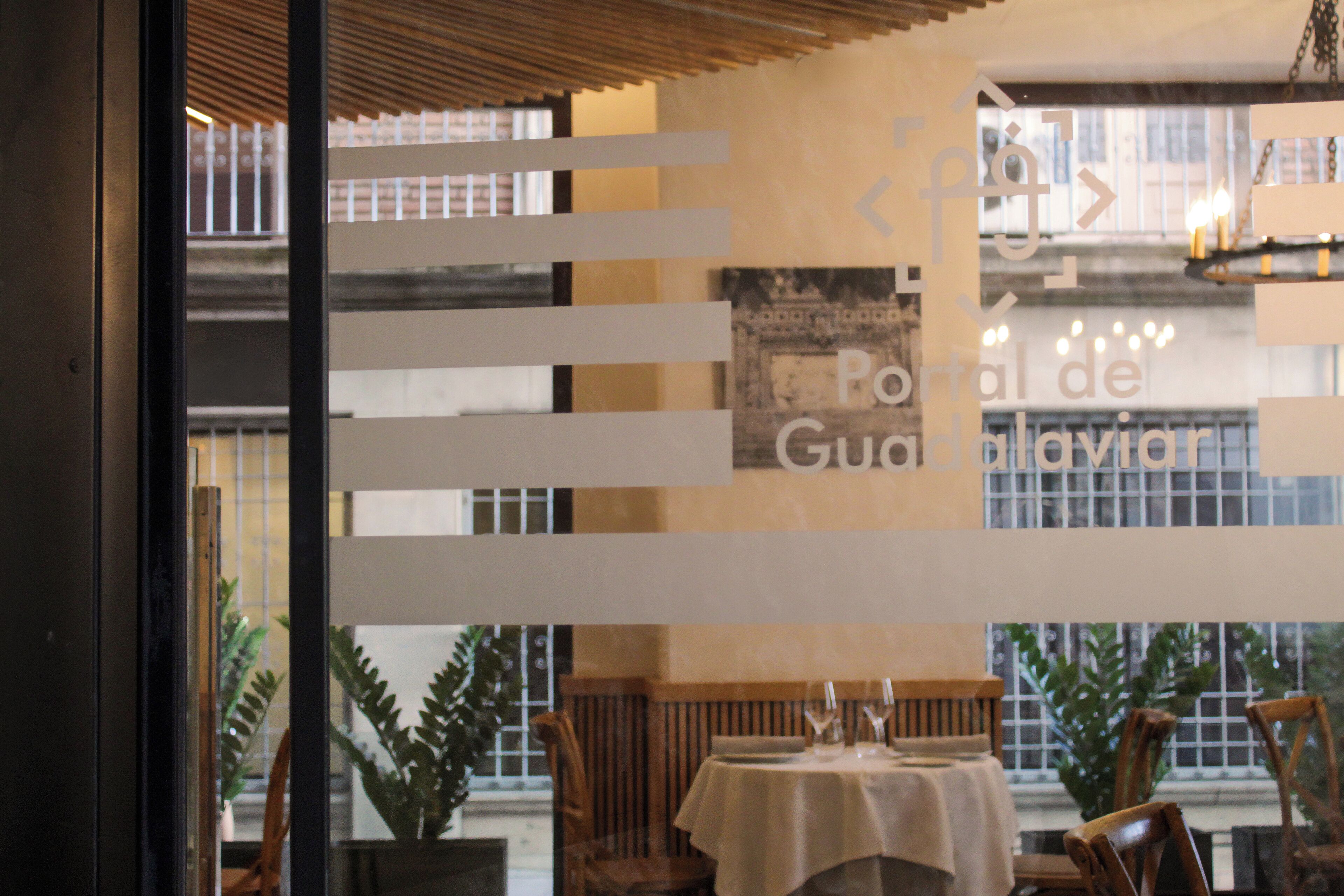 An indoor dining area with tables set for two, wooden chairs, and a view of the outside through a window.