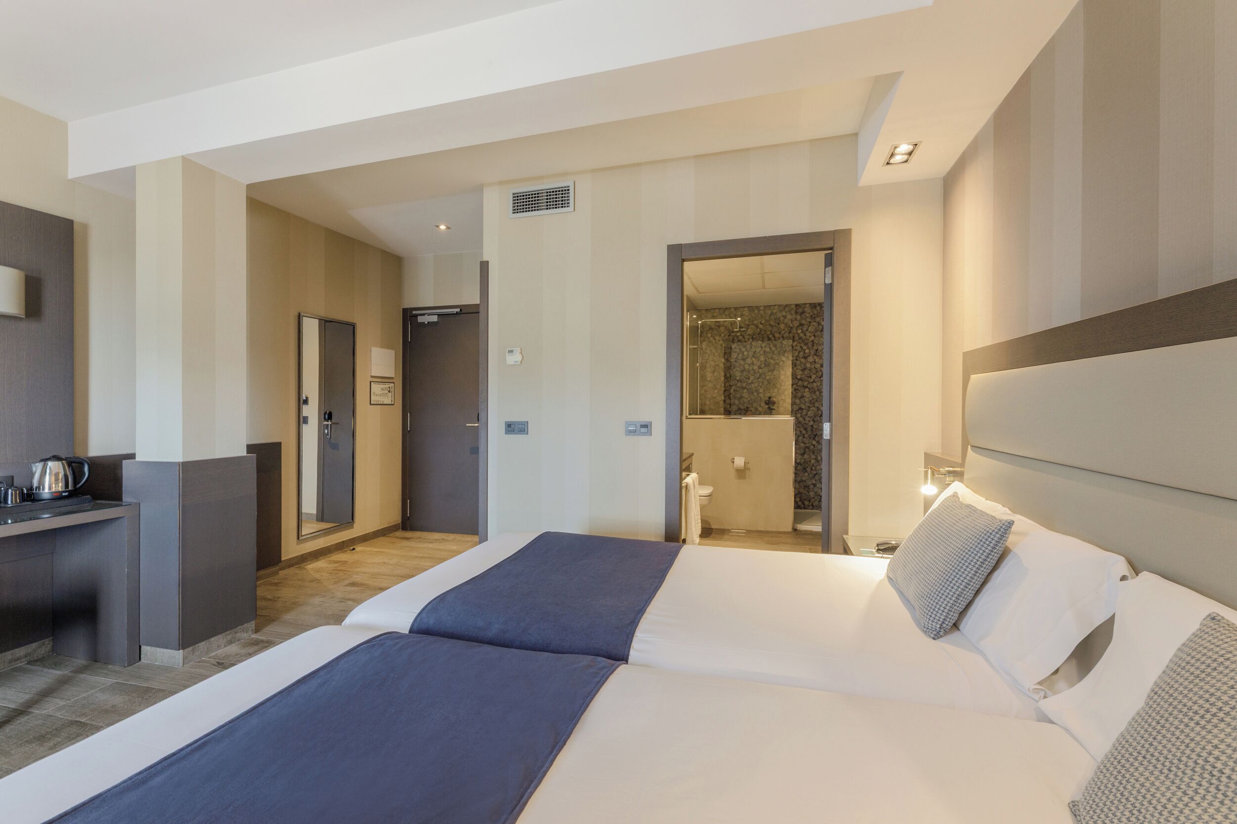 Habitación de hotel con una cama grande, un escritorio con hervidor y un baño visible a través de una puerta abierta.