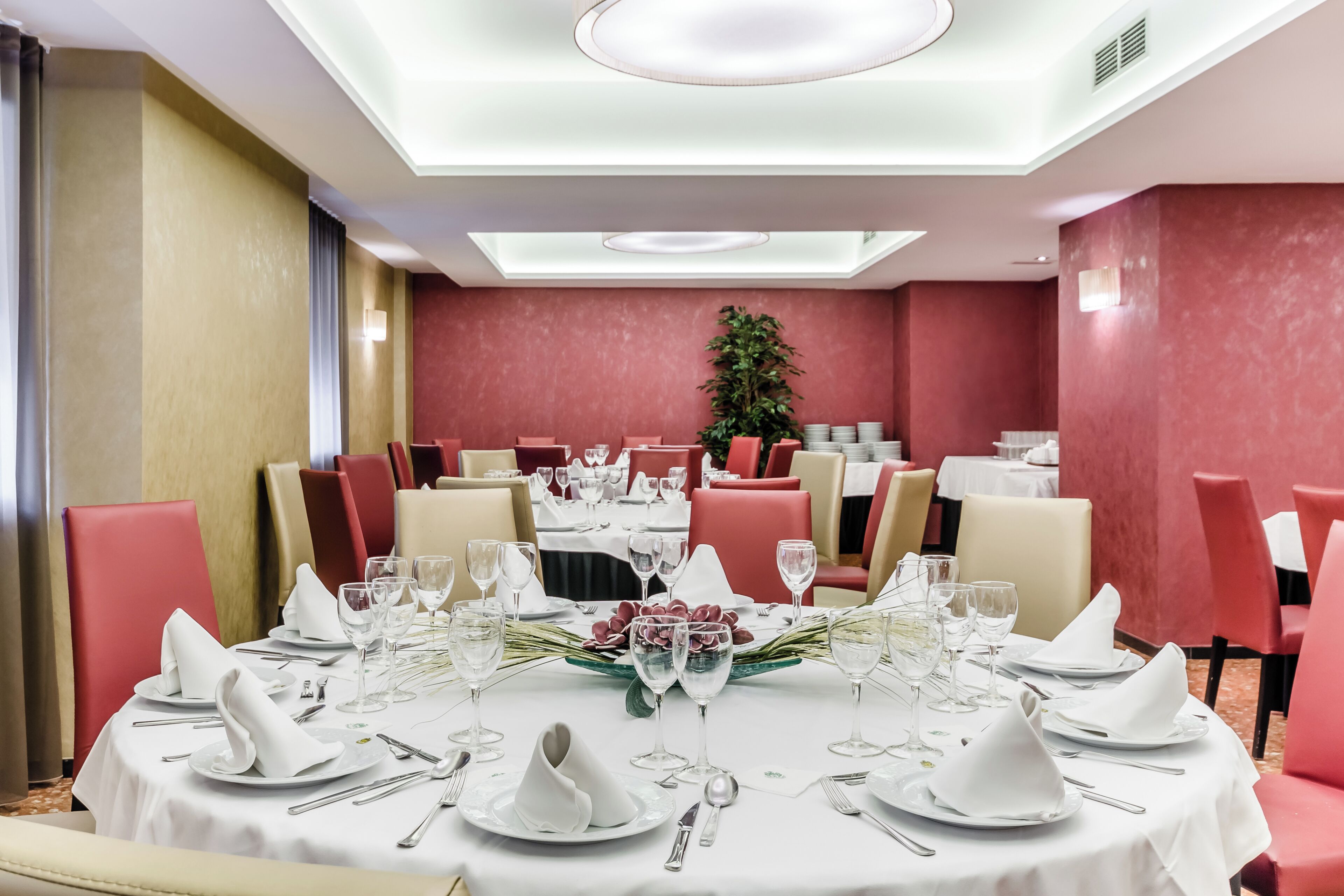 Zona pranzo con tavoli rotondi apparecchiati per un pasto, sedie rosse e beige e una pianta al centro.