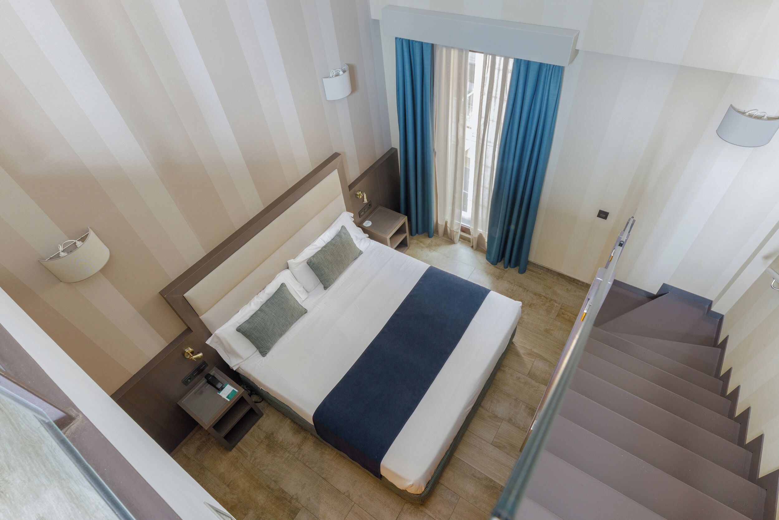Una habitación de hotel con cama, mesita de noche, lámpara, ventana con cortinas y una escalera.