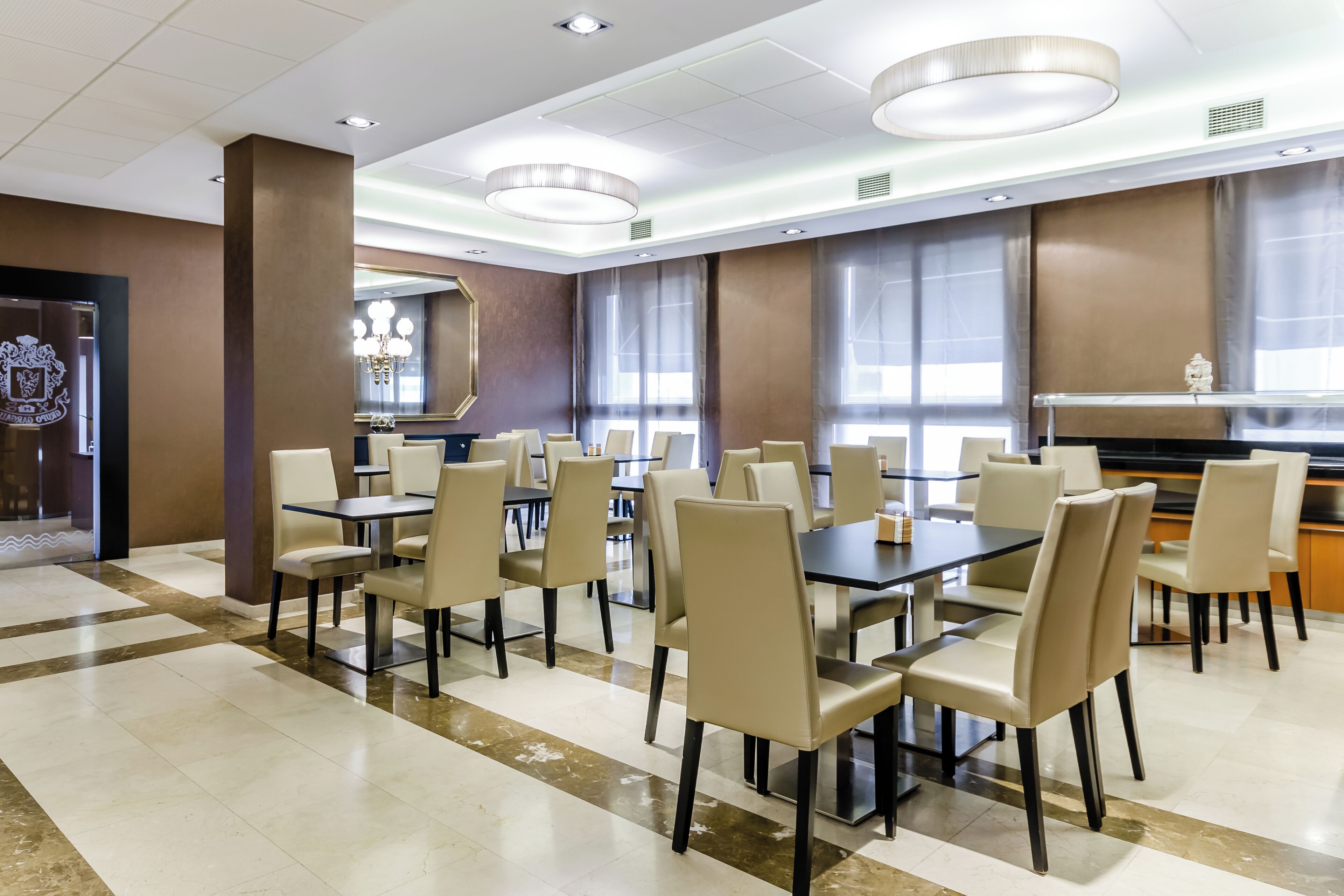 A spacious dining area with multiple tables and chairs, large windows with curtains, and a modern chandelier.