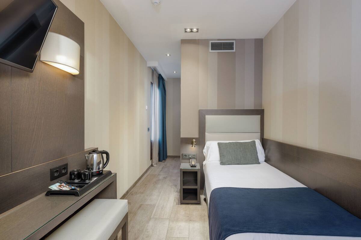 Habitación de hotel con cama, mesita de noche, televisor montado en la pared, una pequeña mesa con hervidor y tazas, y ventana con cortinas.
