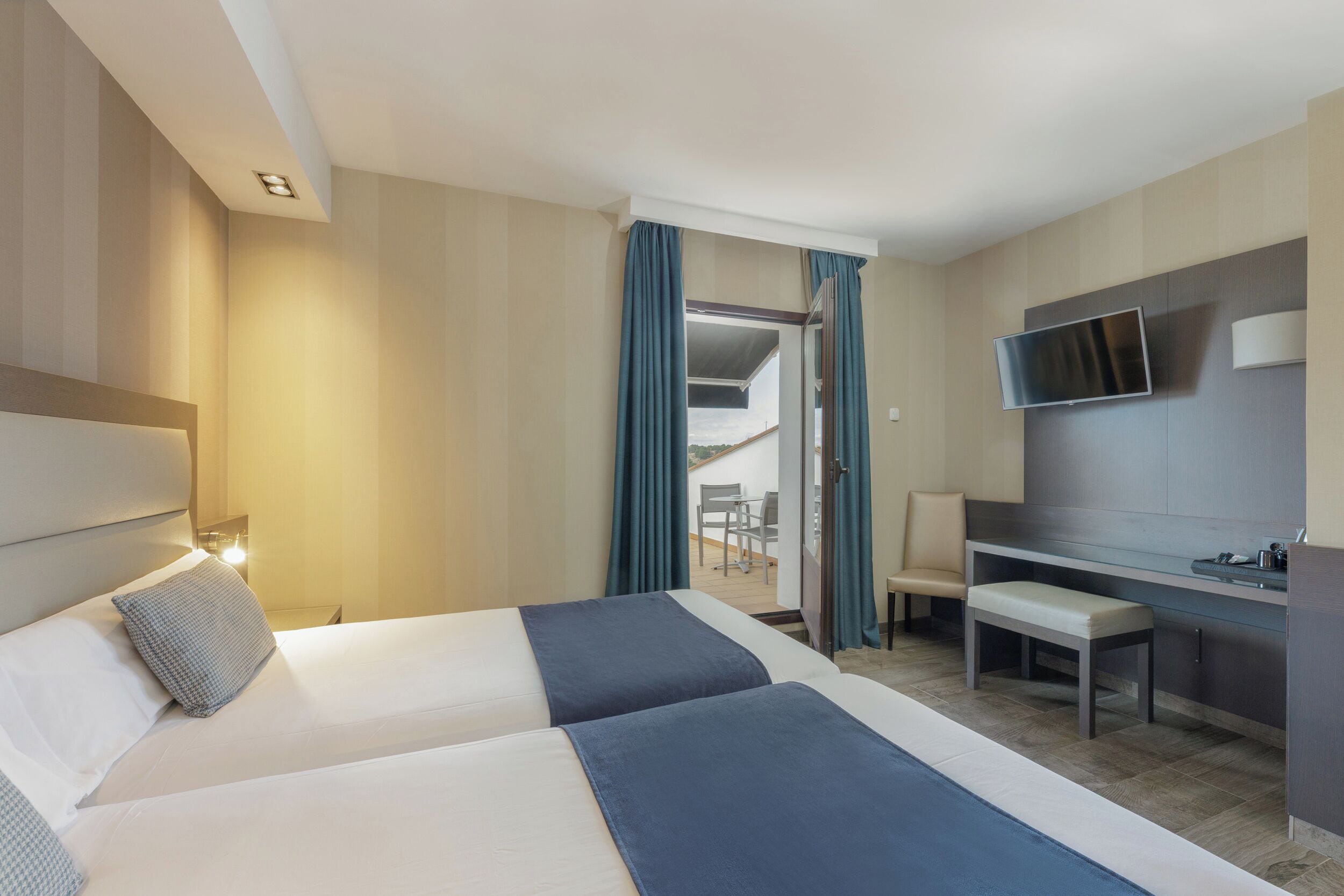 A hotel room with two beds, a desk, a TV, and a balcony with a chair.