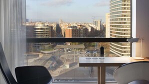 Habitación de hotel moderna con un amplio ventanal que ofrece vistas a la ciudad, un escritorio con lámpara y dos sillones.