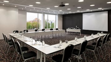 A conference room with a long table, chairs, a projector screen, and a window with a city view.