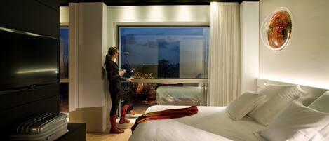 A modern hotel room with a large bed, a TV, and a view of the city at night.