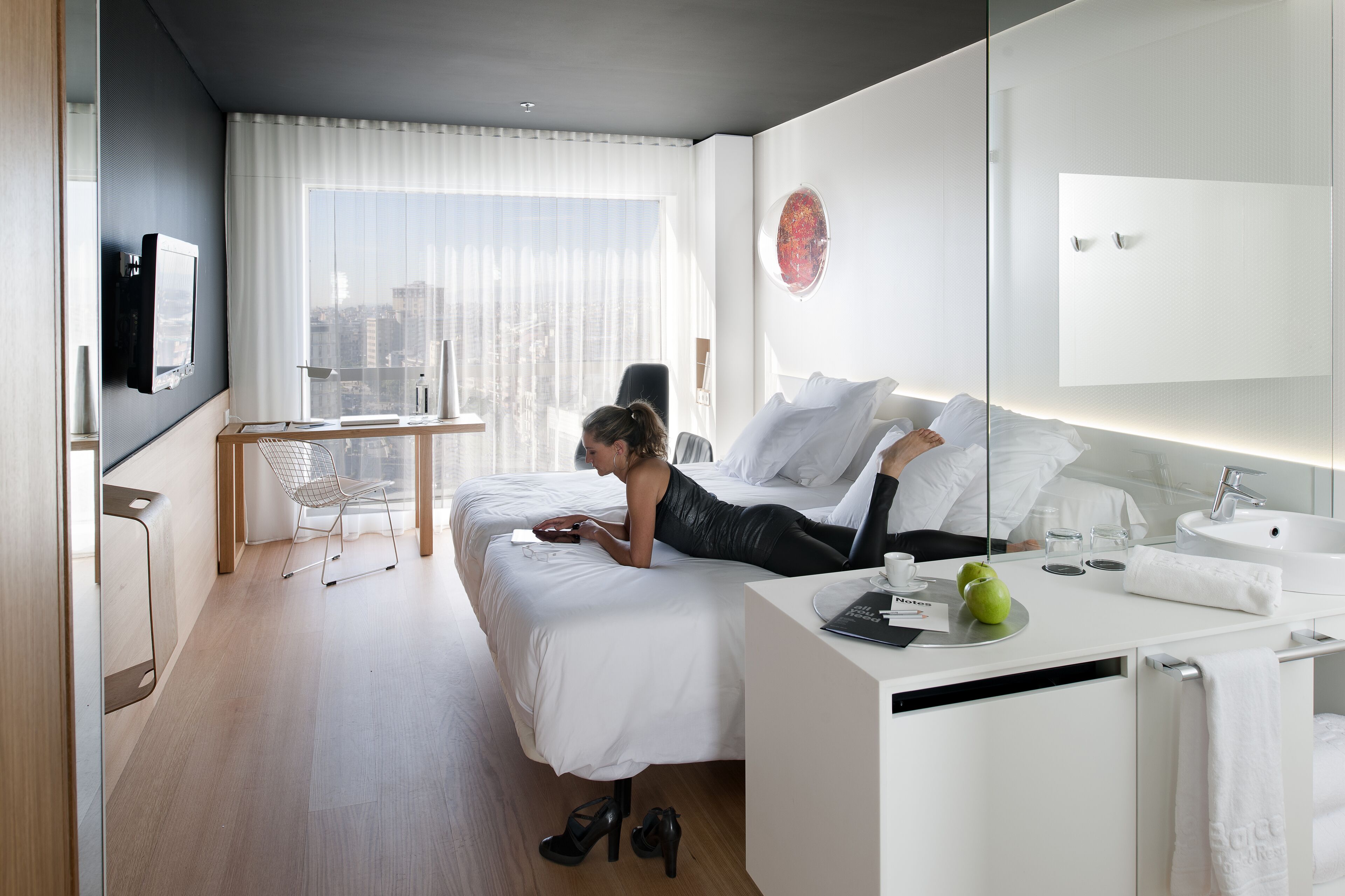 Quarto de hotel moderno com cama, escrivaninha, cadeira, TV e vista para a cidade.