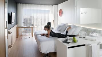 A modern hotel room with a bed, a desk, a chair, a TV, and a city view.