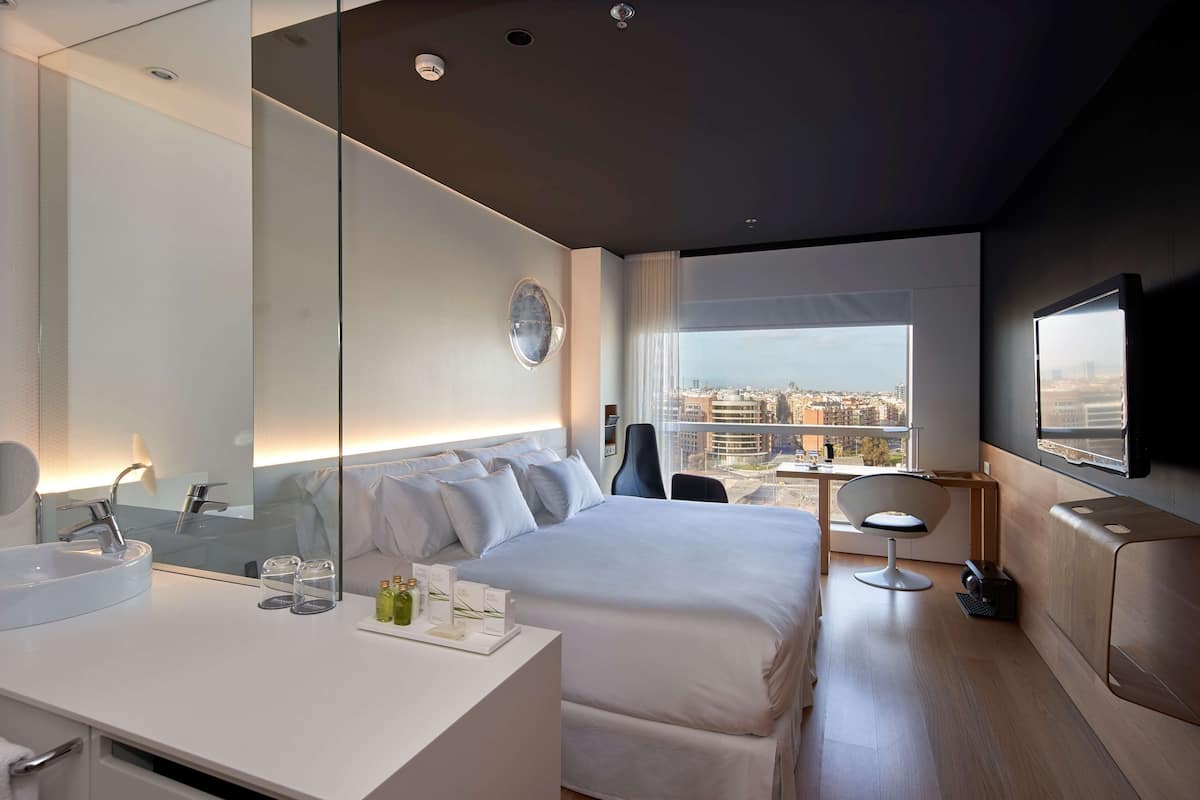 Habitación de hotel moderna con una cama grande, un escritorio con silla, un espejo amplio, un televisor y vistas a la ciudad.