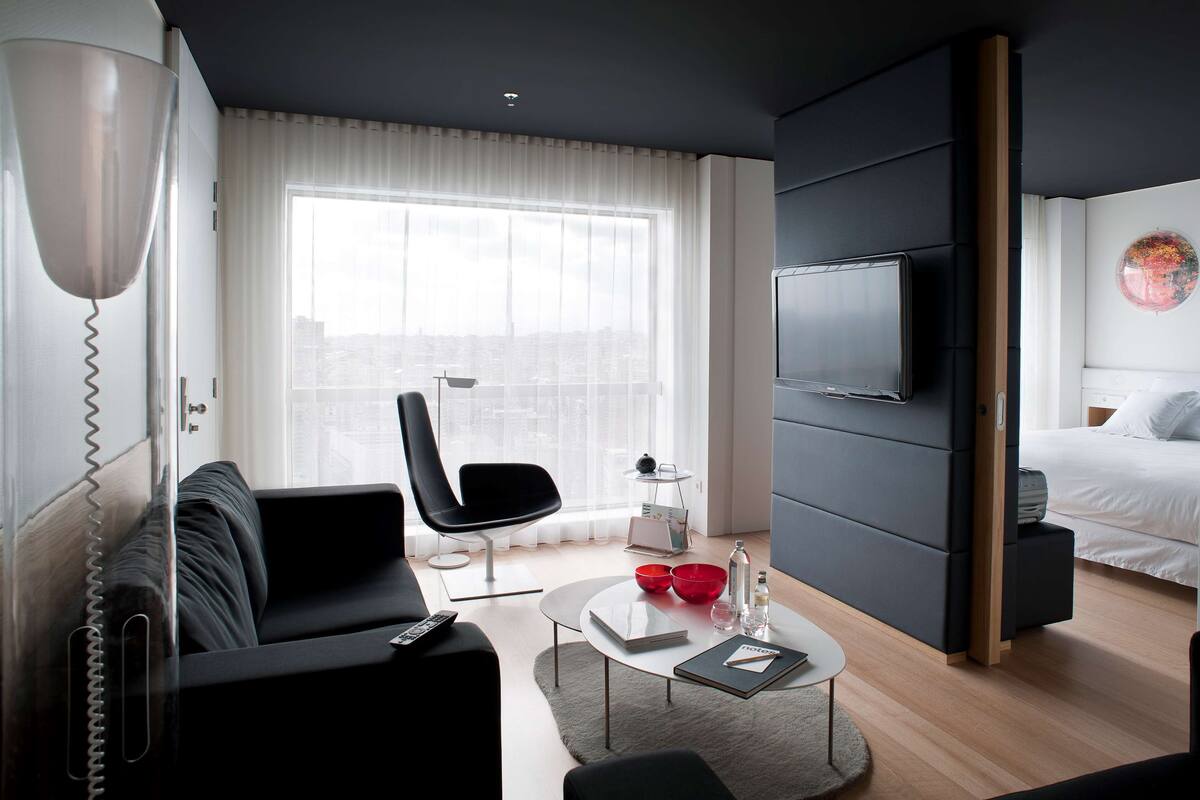 Habitación de hotel moderna con un ventanal grande, un televisor de pantalla plana, una cama, un sofá y una mesa de centro con control remoto.