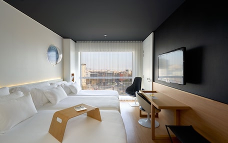 A modern hotel room with two beds, a desk, a chair, a TV, and a view of the city.