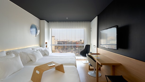 Habitación de hotel moderna con dos camas, un escritorio, una silla, un televisor y vistas a la ciudad.