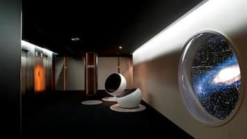 Un moderno vestíbulo de hotel con un amplio ventanal circular que muestra un cielo estrellado, un área de descanso con sillas originales y un ascensor elegante.