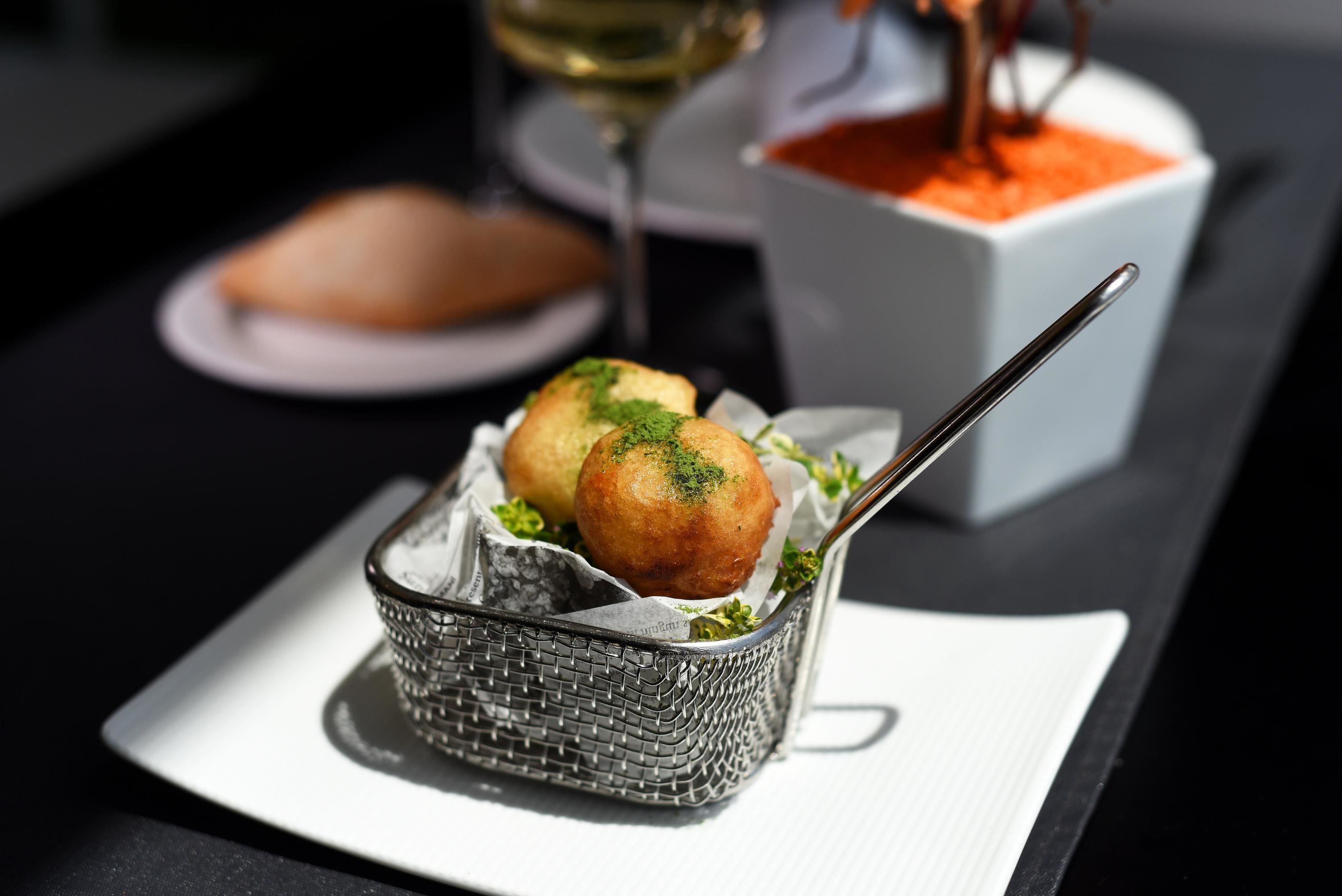 A basket of food items on a table with a wine glass and a bowl of sauce in the background.