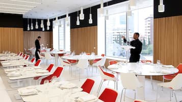 A modern dining area with round tables and red chairs, large windows with a city view, and a waiter attending to guests.