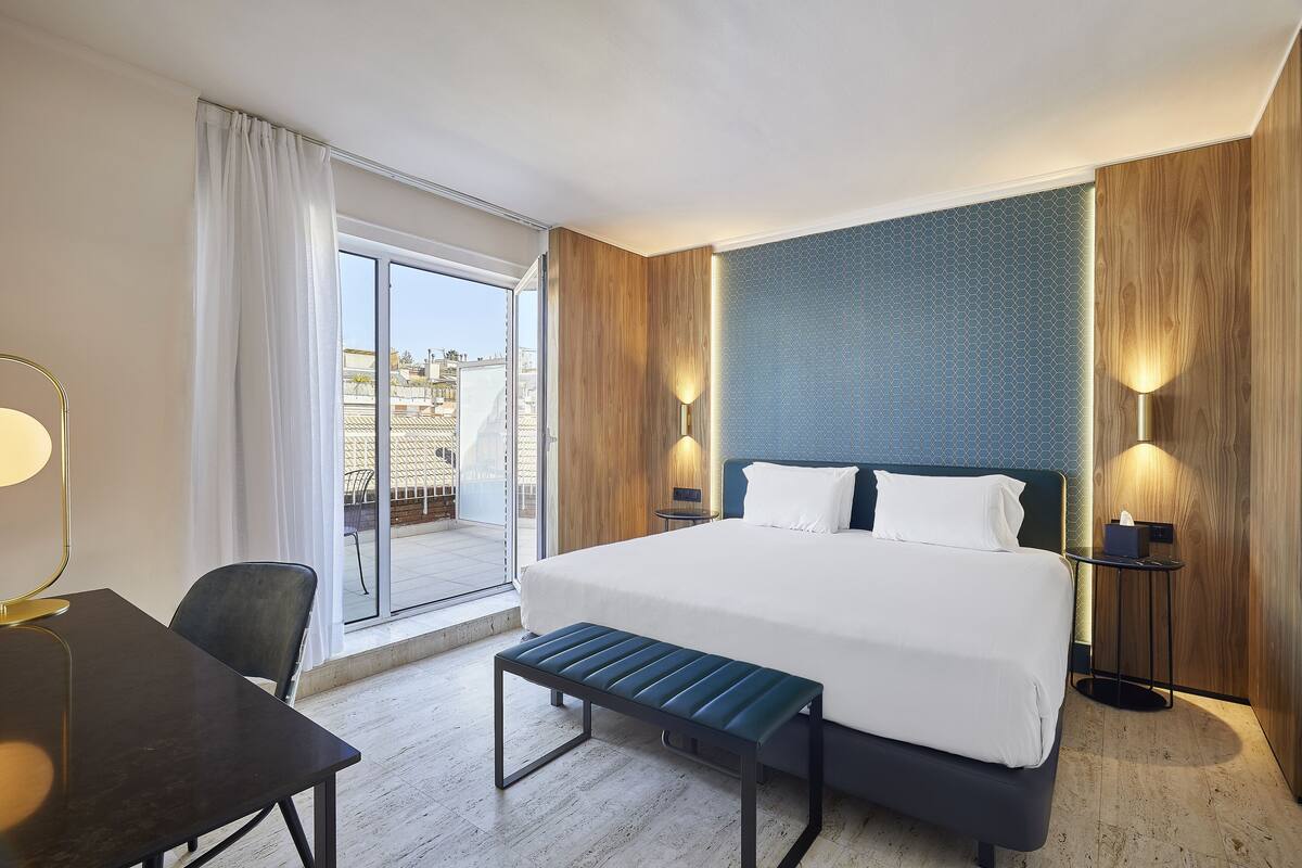 A hotel room with a large bed, a desk, and a bench. There is a window with curtains, a lamp, and a view of buildings outside.