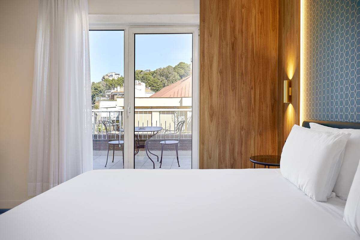 A hotel room with a large bed, a wooden accent wall, and a view of a balcony with outdoor furniture.
