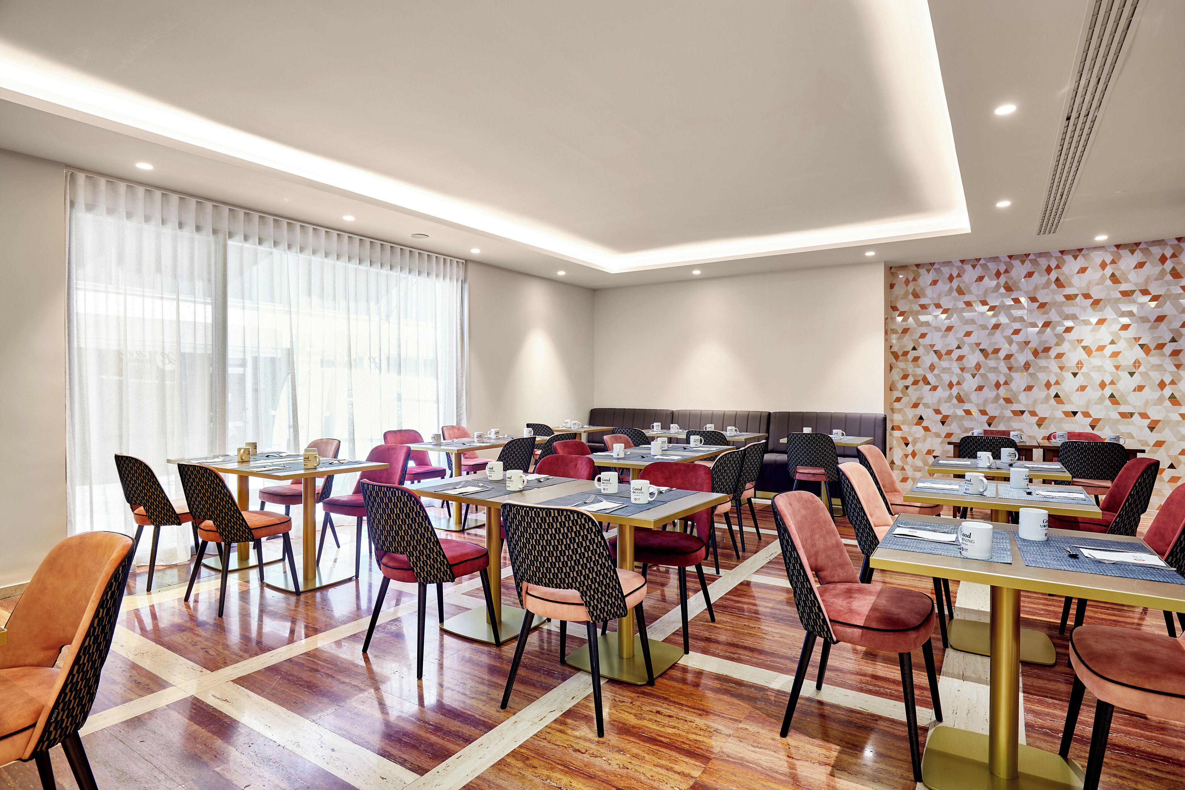 Una sala de conferencias moderna con varias mesas y sillas, ventanas amplias con cortinas translúcidas y un muro con diseño.
