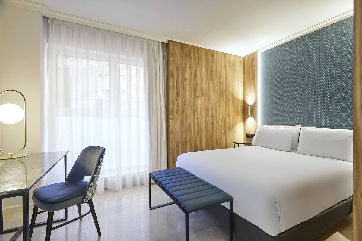 A hotel room with a bed, a desk, a chair, and a bench.