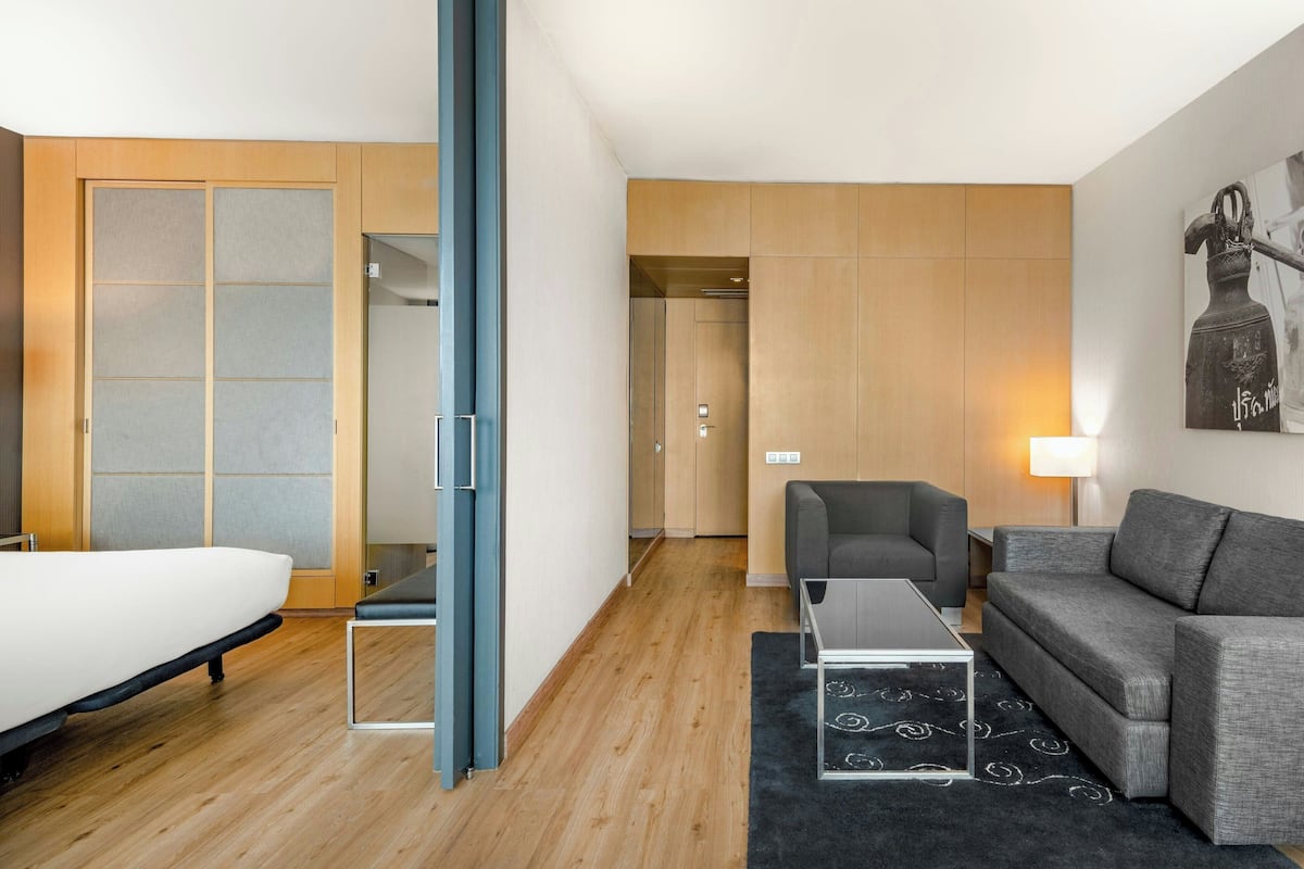 A modern hotel room with a bed, a grey sofa, a small table, and a black rug. There is a wooden wardrobe and a door leading to a hallway.