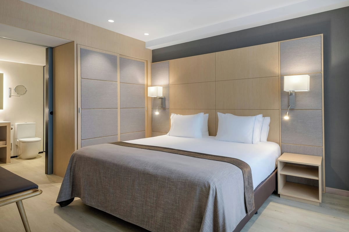 A modern hotel room with a large bed, bedside tables, a bedside lamp, a wardrobe, and a small desk with a chair.