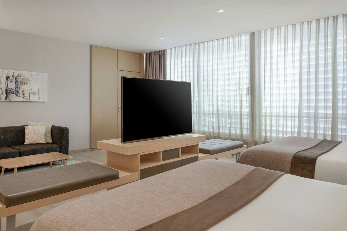 A modern hotel room with a flat-screen TV, a sofa, a bed, and a large window with sheer curtains.