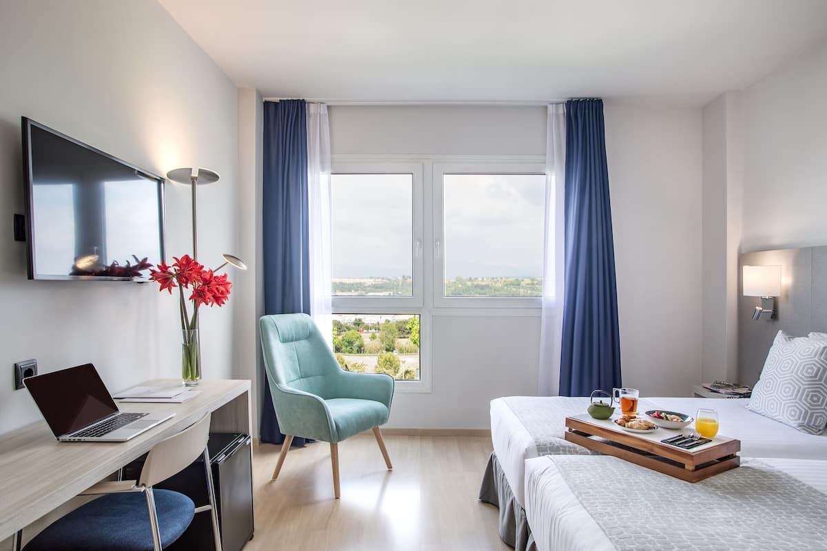 Quarto de hotel com escrivaninha, cadeira, cama, bandeja de café da manhã e televisão na parede.