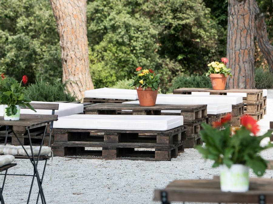 Mobiliario exterior con mesas y bancos de madera, rodeado de plantas en macetas y árboles.