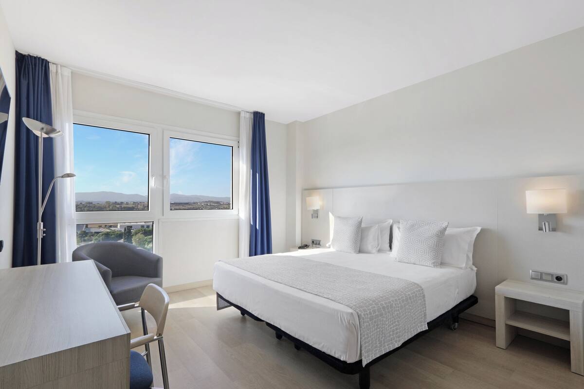 Quarto de hotel com uma cama grande, uma escrivaninha e uma cadeira. Há duas janelas com cortinas e vista para o exterior.