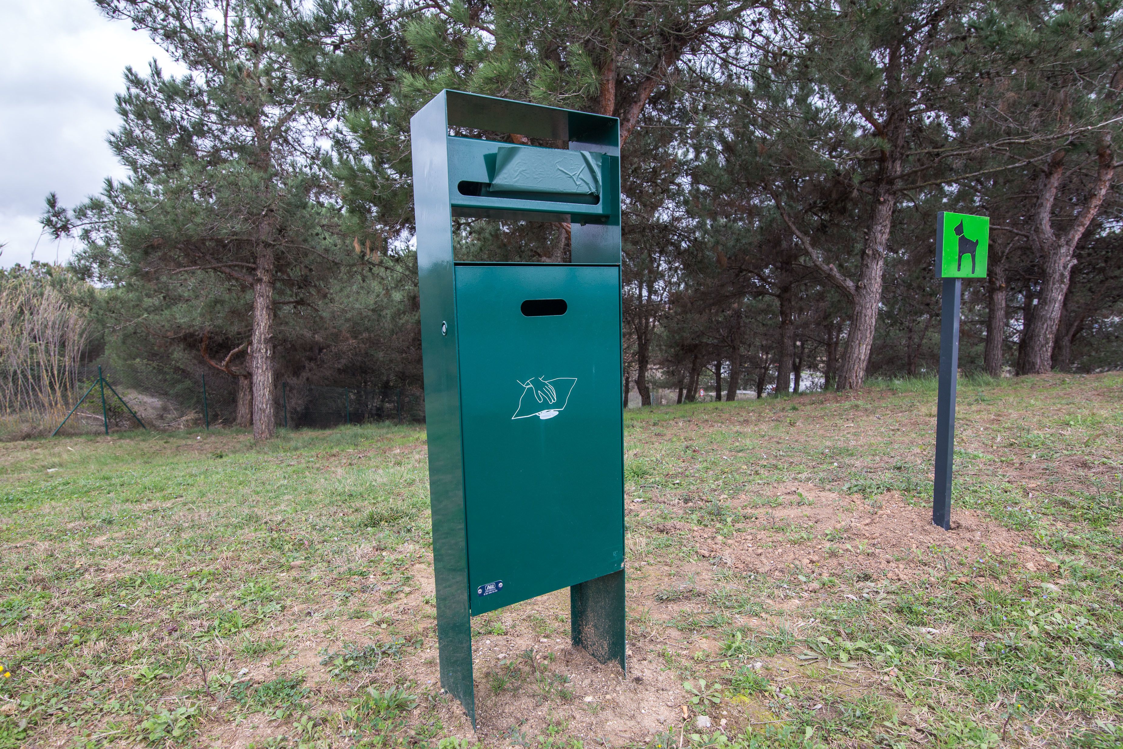 A green outdoor trash bin with a recycling symbol, located in a grassy area with trees in the background.