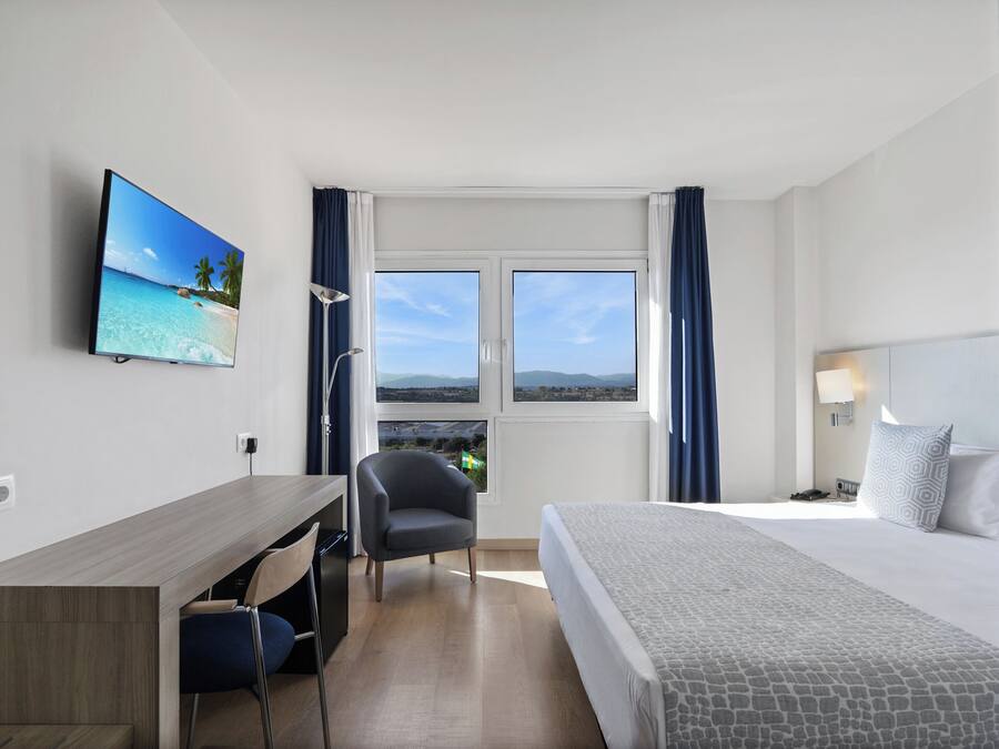 Habitación de hotel con cama, escritorio, silla, televisor y ventana con vista a la playa y las montañas.