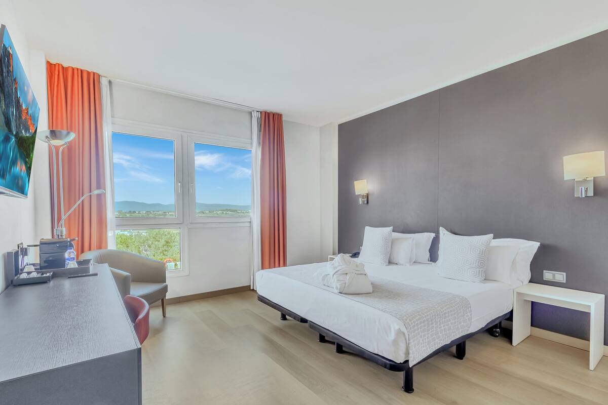 Quarto de hotel com cama, mesa de cabeceira, escrivaninha, cadeira e uma grande janela com cortinas.
