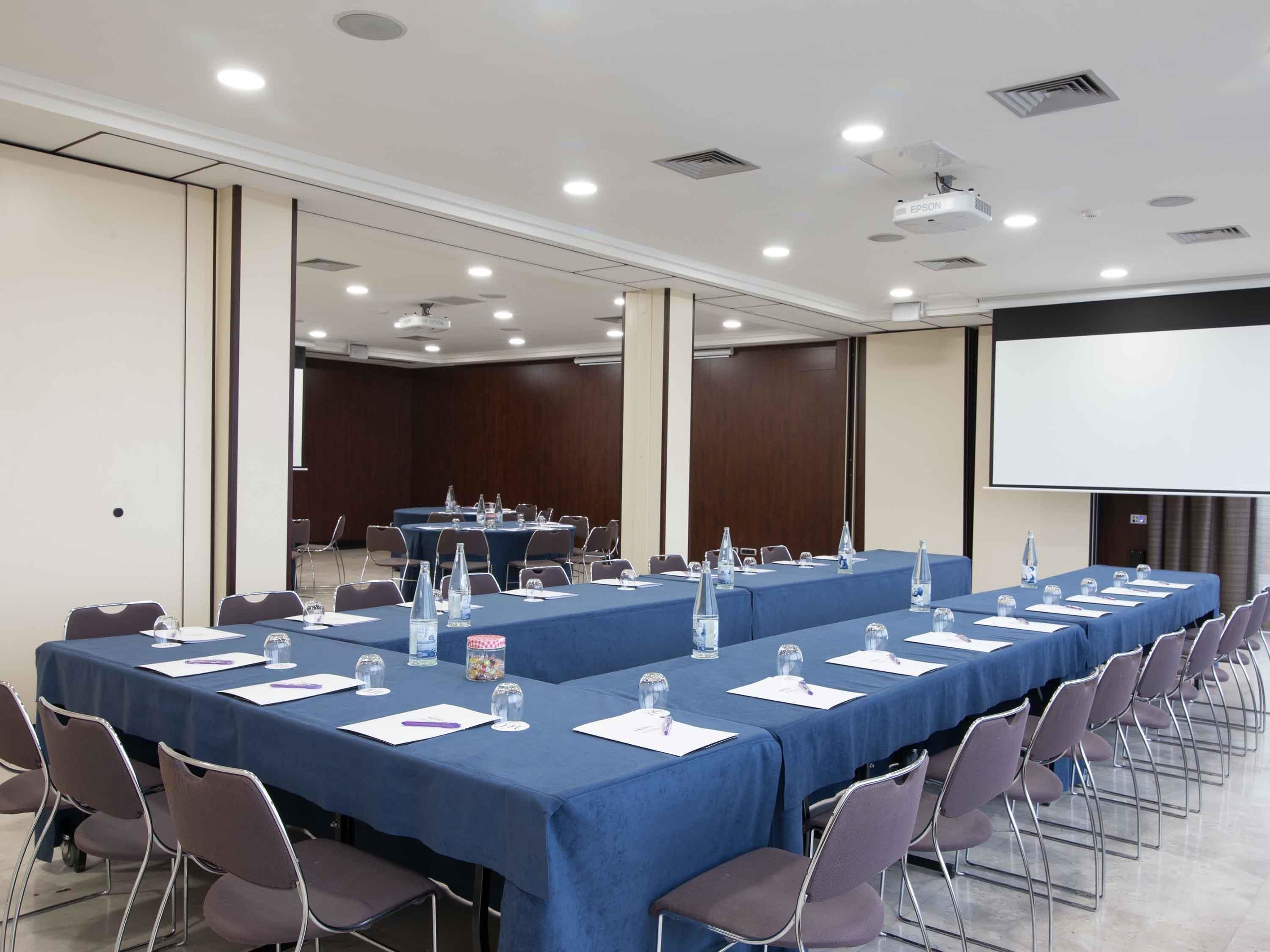 Un salón de conferencias con mesas largas, sillas y una pantalla para proyector.