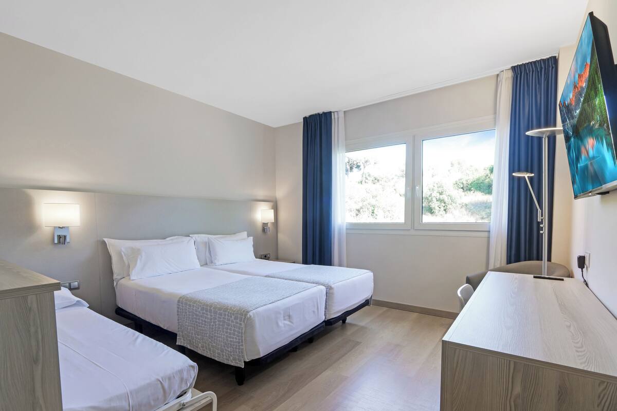 Quarto de hotel com duas camas, uma escrivaninha, uma cadeira, televisão e janela com cortinas.